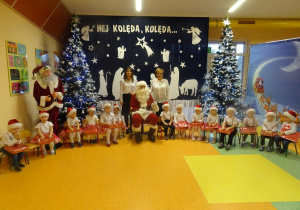 Pani dyrektor Maria Królikowska, pani Ewelina Cicha i dzieci zebrane wokół Mikołaja pozują na tle dekoracji świątecznej.